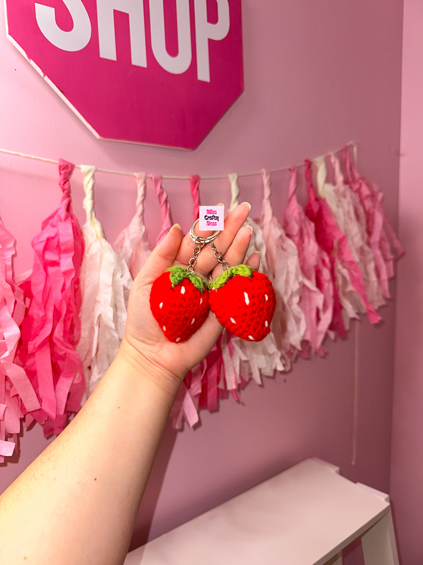 Hand holding two red strawberry-shaped keychains against a pink wall with decorative tassels and a 'Shop' sign.