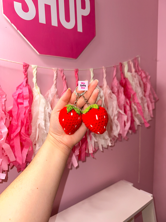 Hand holding two red strawberry-shaped keychains against a pink wall with decorative tassels and a 'Shop' sign.