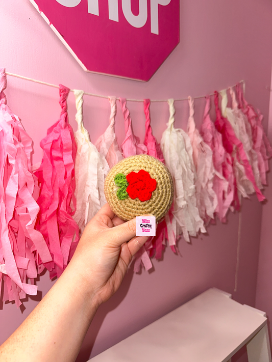 Hand holding a crocheted round candy with a red flower in the middle against a pink wall with tassels