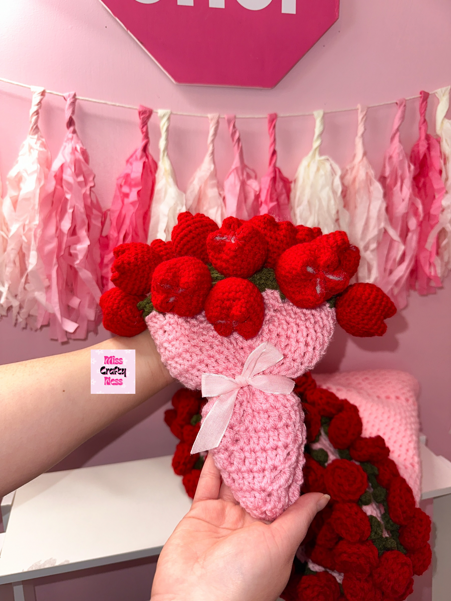 Handmade crochet bouquet of red tulips with a pink base held by a person against a pink wall with decorative ribbons.