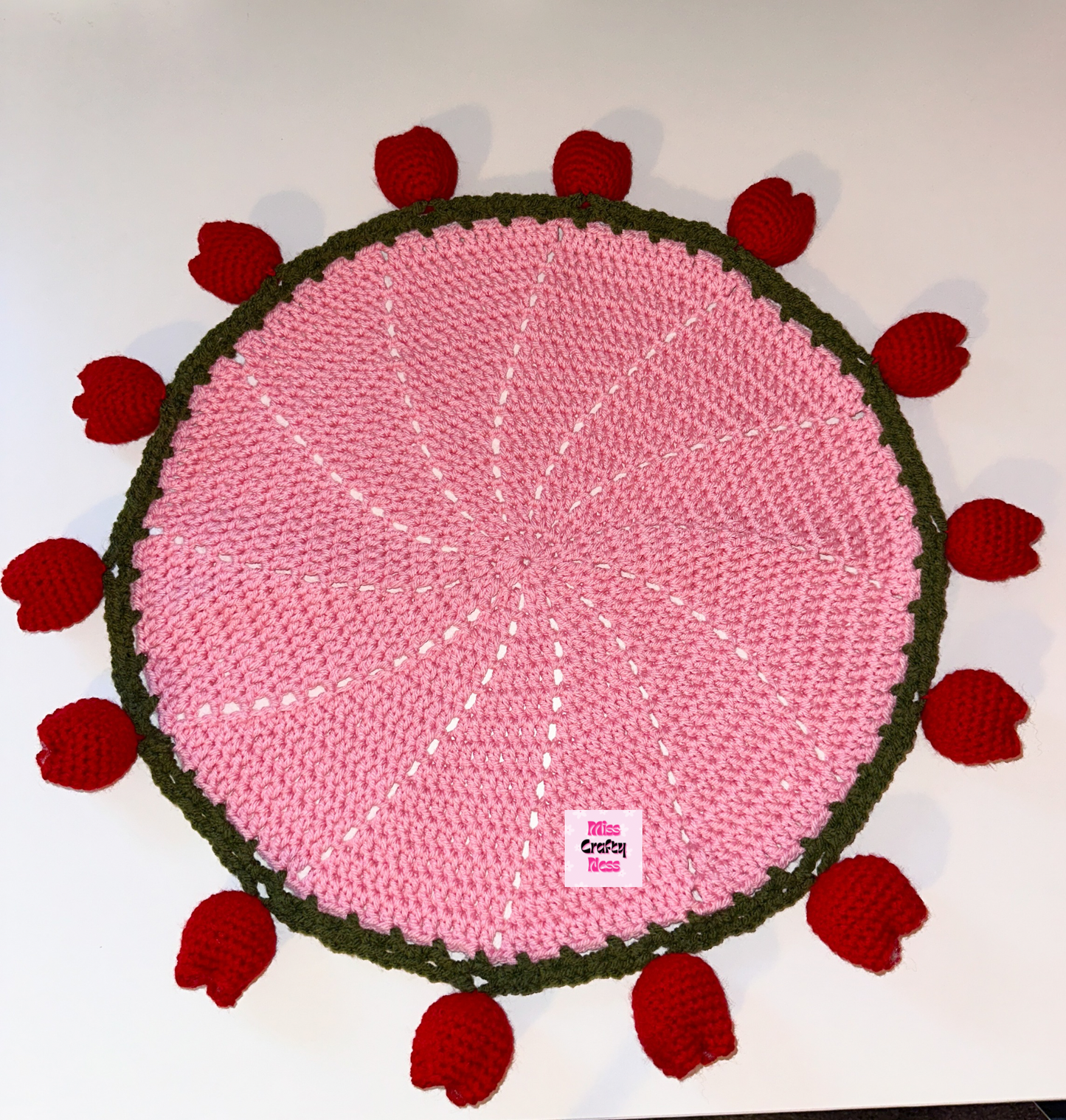Round pink crocheted blanket with green border and red tulip flower bulbs on a white background