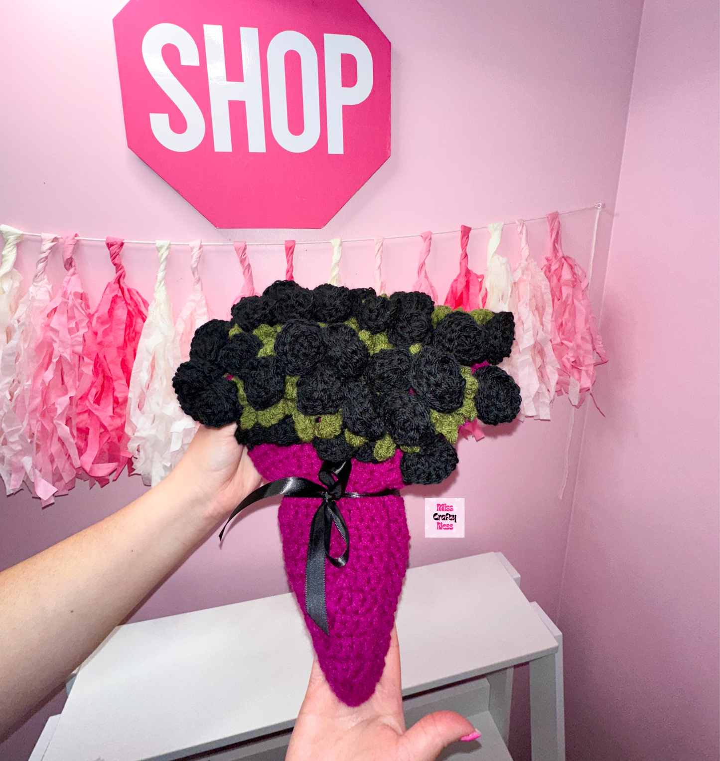 Crocheted bouquet blanket, base is violet with a black ribbon bow. Topped with black roses, held by a hand against a pink wall with decorative tassels and a sign that says “SHOP”.