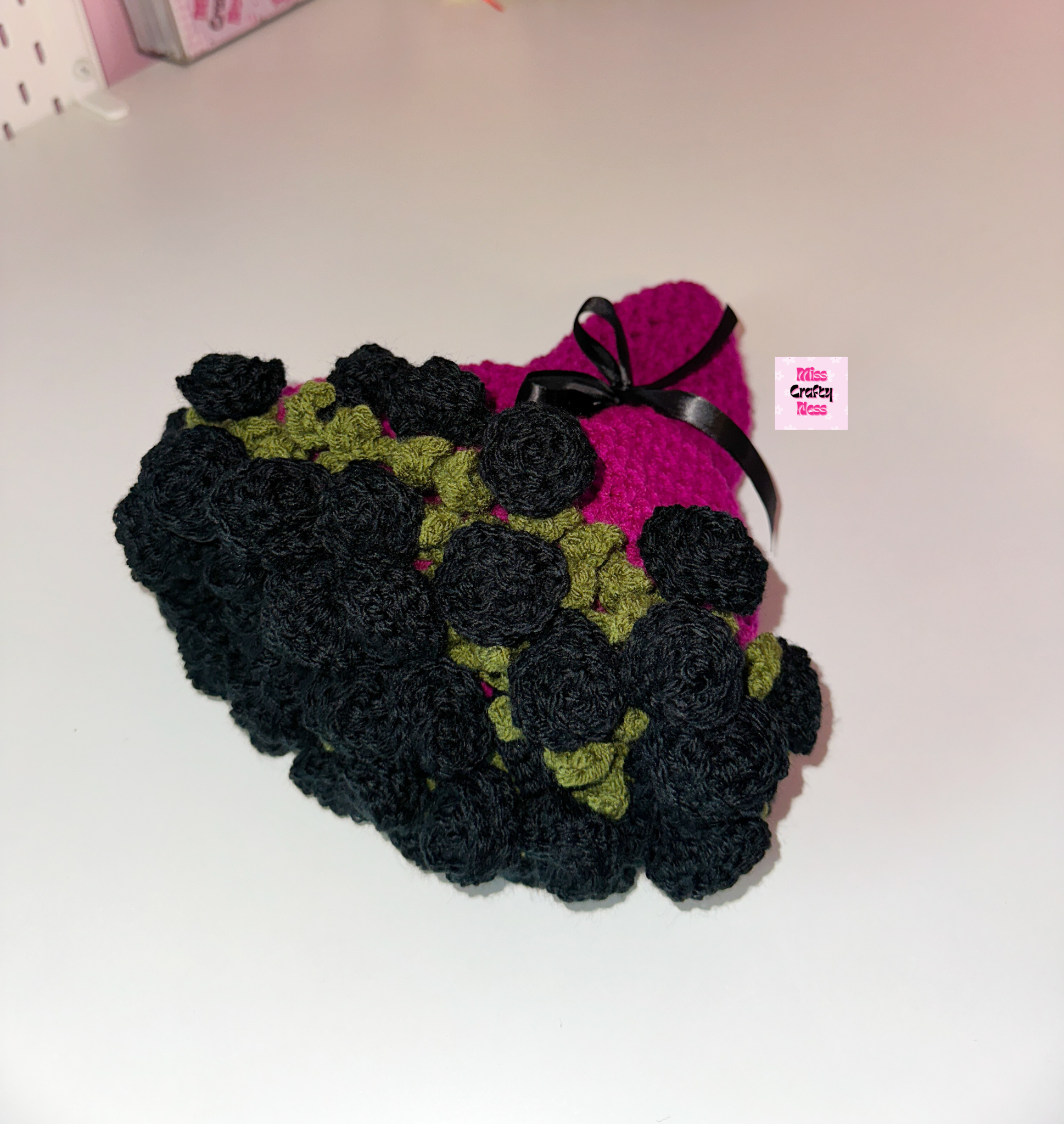 Crocheted blanket bouquet with black roses, green leaves and a violet base wrapped in a black bow on a white surface with a small logo label.
