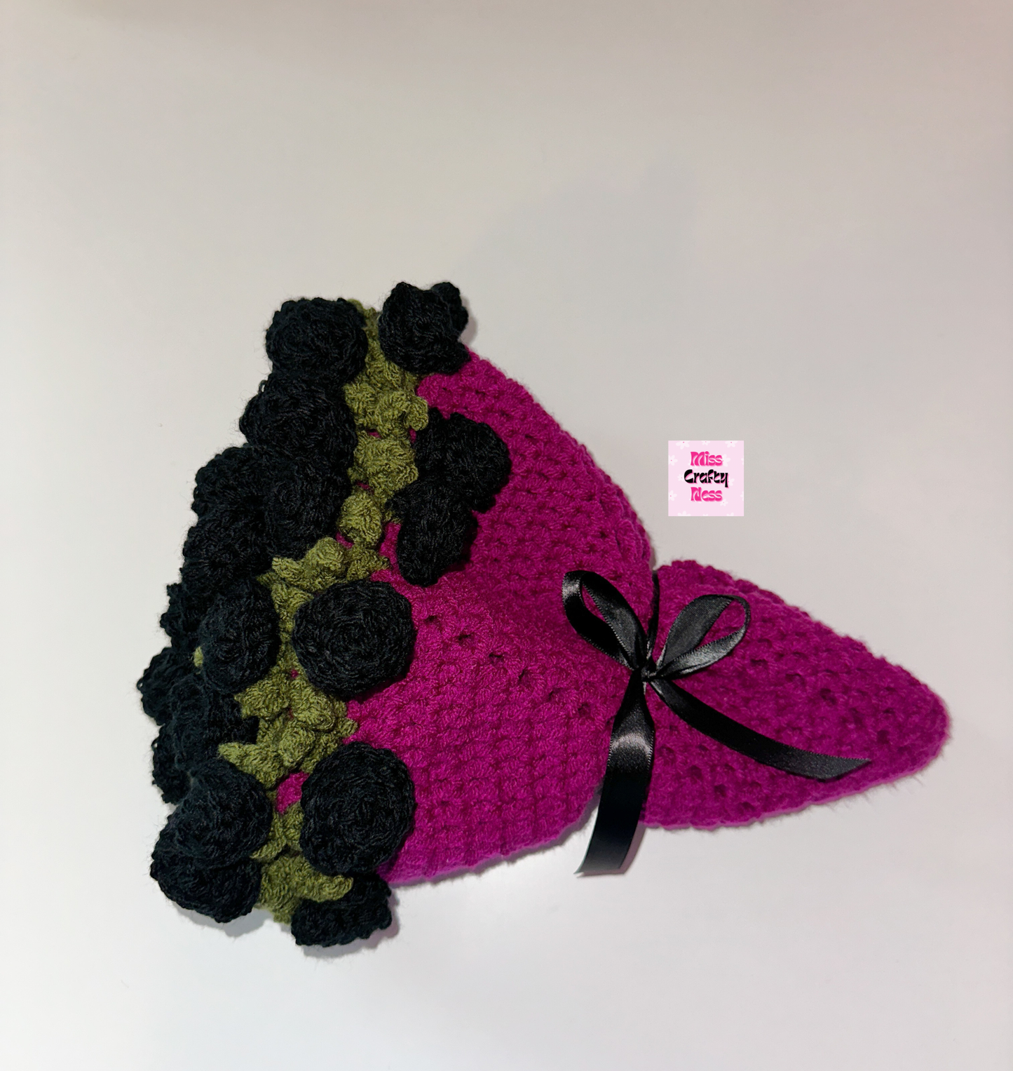 Crocheted blanket bouquet with black roses, green leaves and a violet base wrapped in a black bow on a white surface with a small logo label.