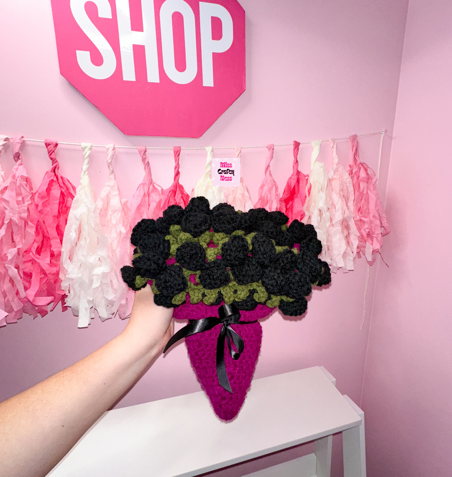 Crocheted bouquet blanket, base is violet with a black ribbon bow. Topped with black roses, held by a hand against a pink wall with decorative tassels and a sign that says “SHOP”.