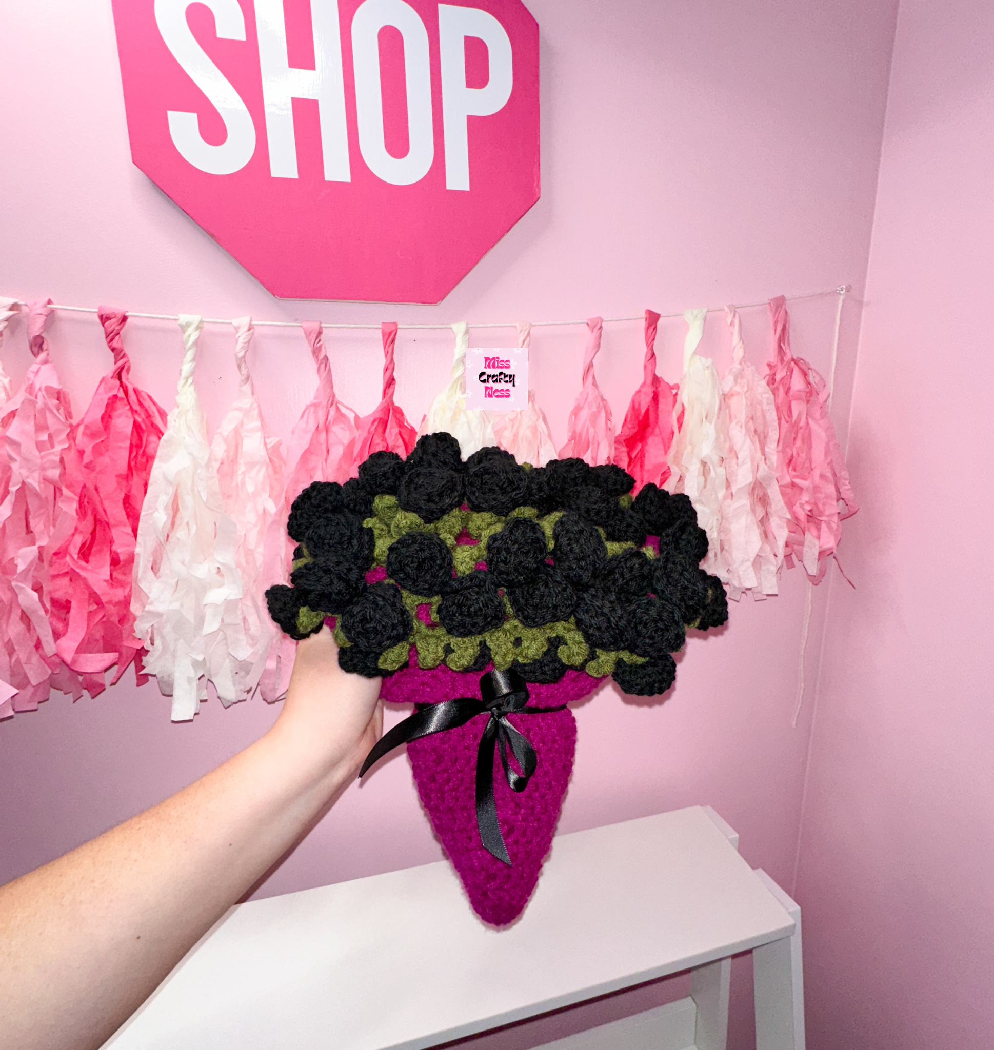 Crocheted bouquet blanket, base is violet with a black ribbon bow. Topped with black roses, held by a hand against a pink wall with decorative tassels and a sign that says “SHOP”.