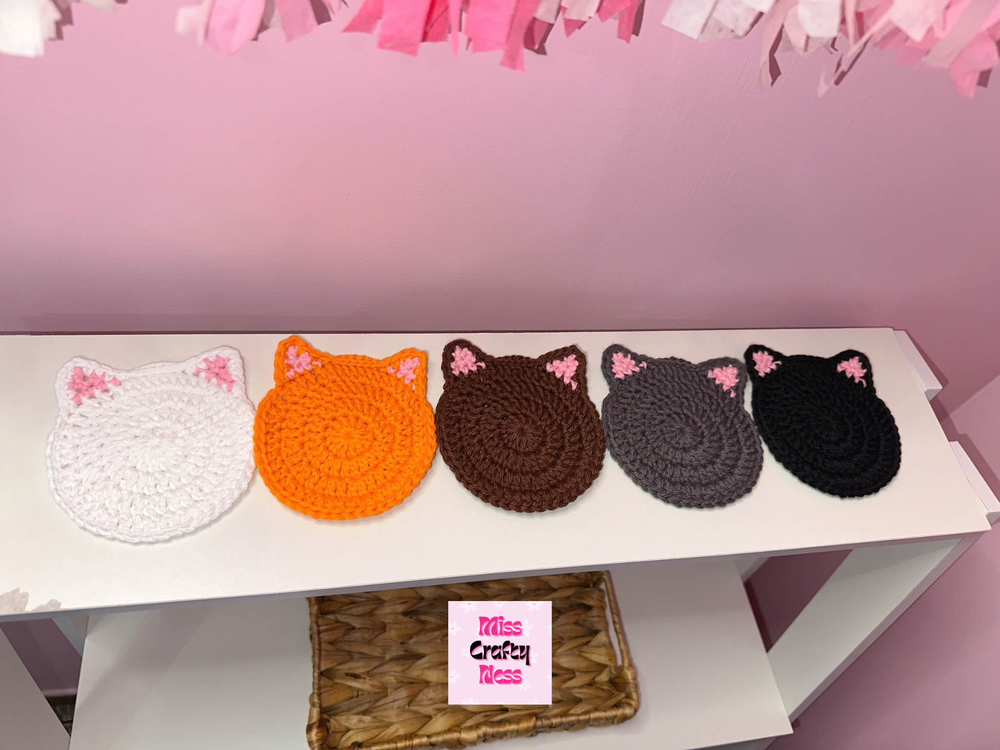 Five crocheted cat-shaped coasters in various colors on a white surface with a pink background.