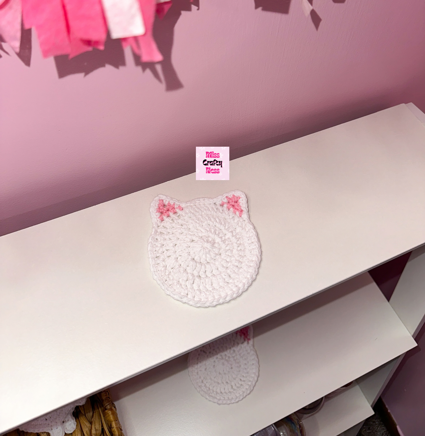 White cat-shaped coaster with pink ears on a white surface against a pink wall.