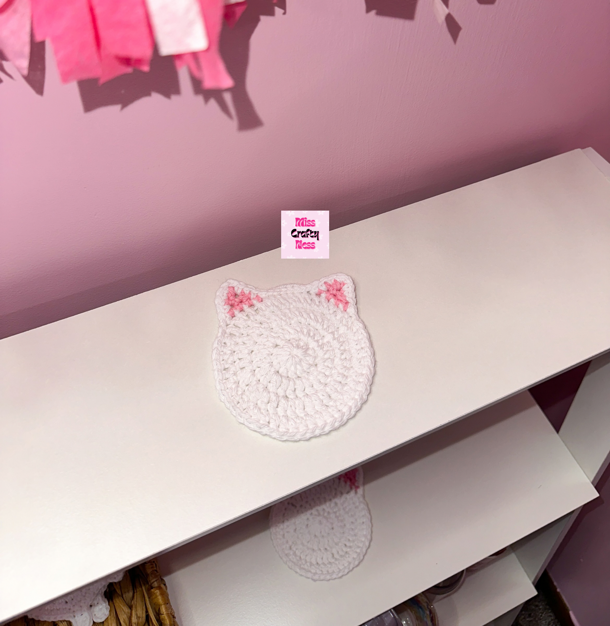 White cat-shaped coaster with pink ears on a white surface against a pink wall.