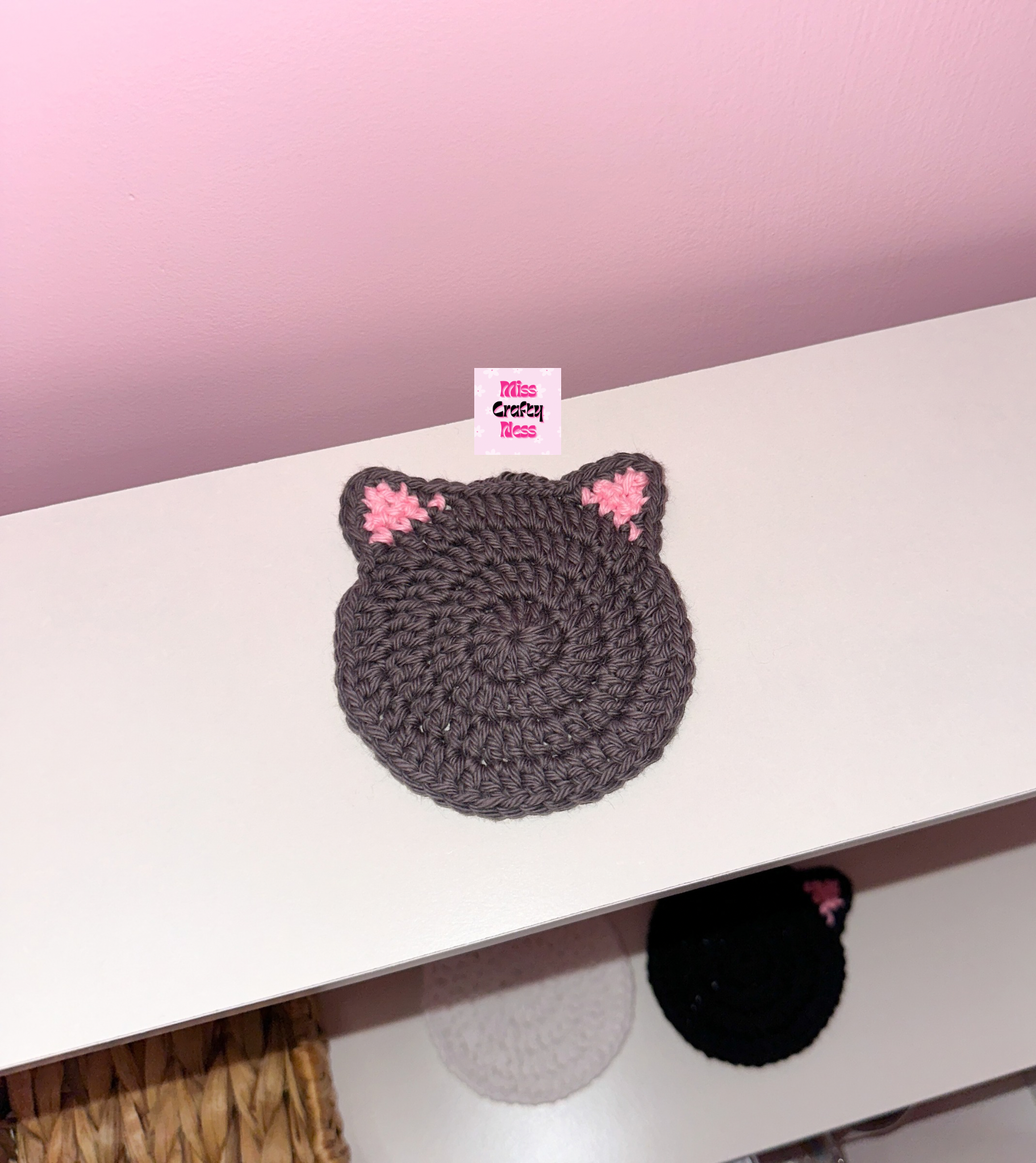 Crocheted cat-shaped coaster on a white surface against a pink background.