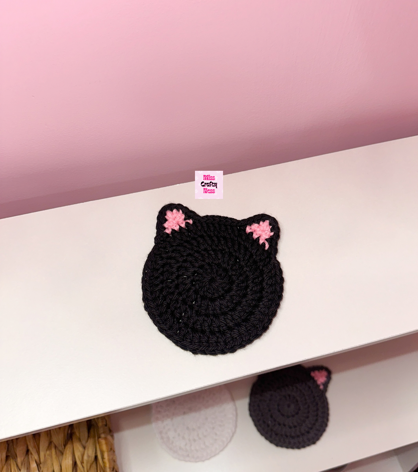 Black crocheted cat-shaped coaster on a white surface with a pink background.