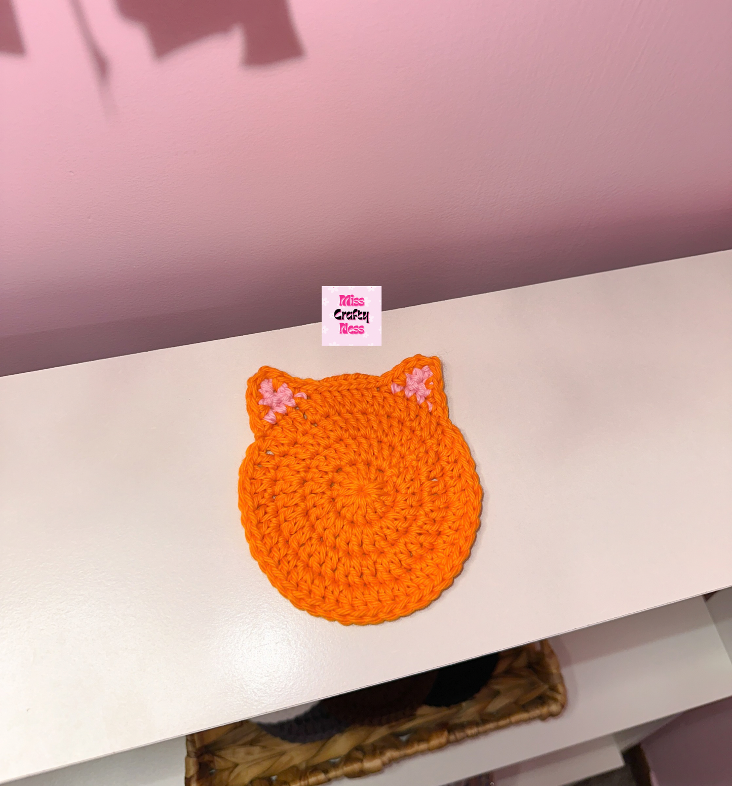 Orange crocheted cat-shaped coaster on a white surface with a pink background