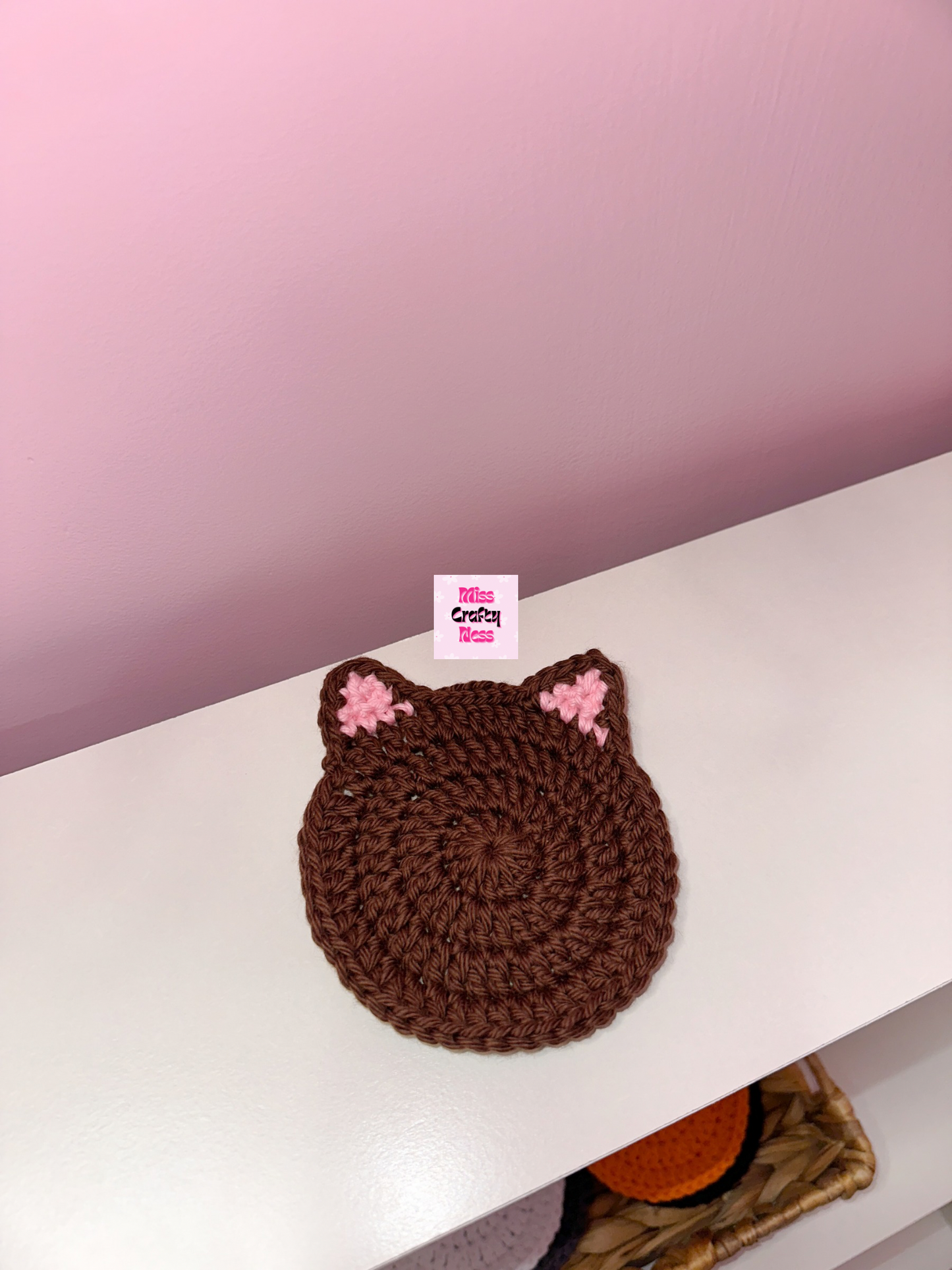 Brown crocheted cat-shaped coaster on a white surface against a pink background.
