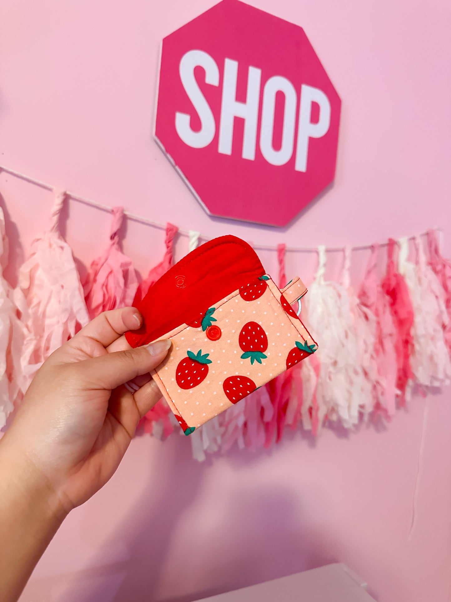 Hand holding a small wallet with strawberry pattern in front of a pink 'SHOP' sign with a pink wall and tassels.